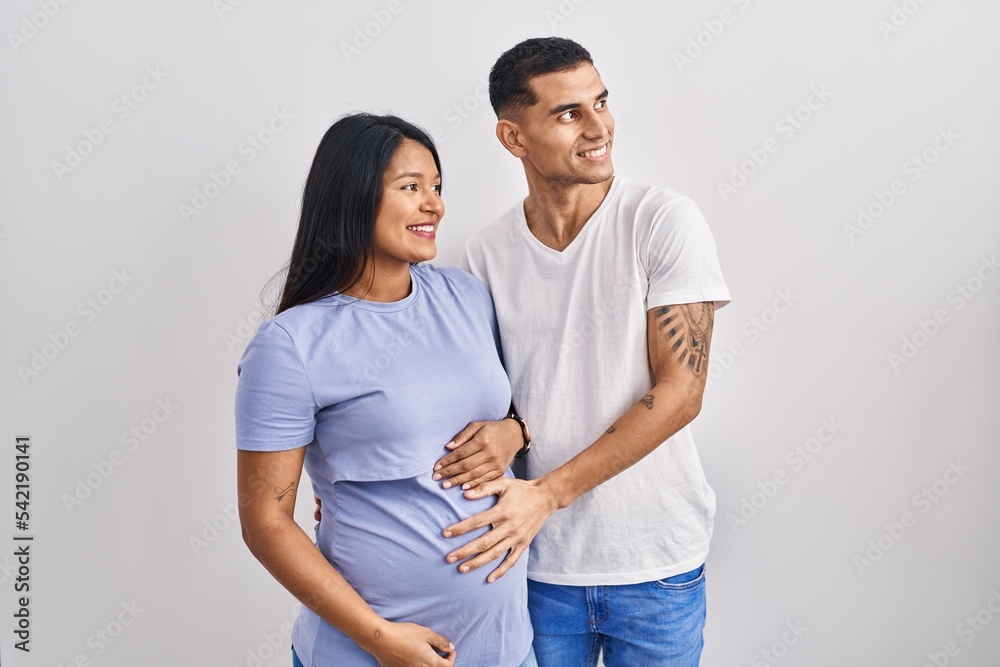 Young hispanic couple expecting a baby standing over background looking away to side with smile on face, natural expression. laughing confident. Young hispanic couple expecting a baby standing over background looking away to side with smile on face, natural expression. laughing confident.