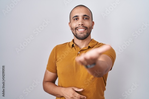 Hispanic man with beard standing over white background smiling cheerful offering palm hand giving assistance and acceptance.