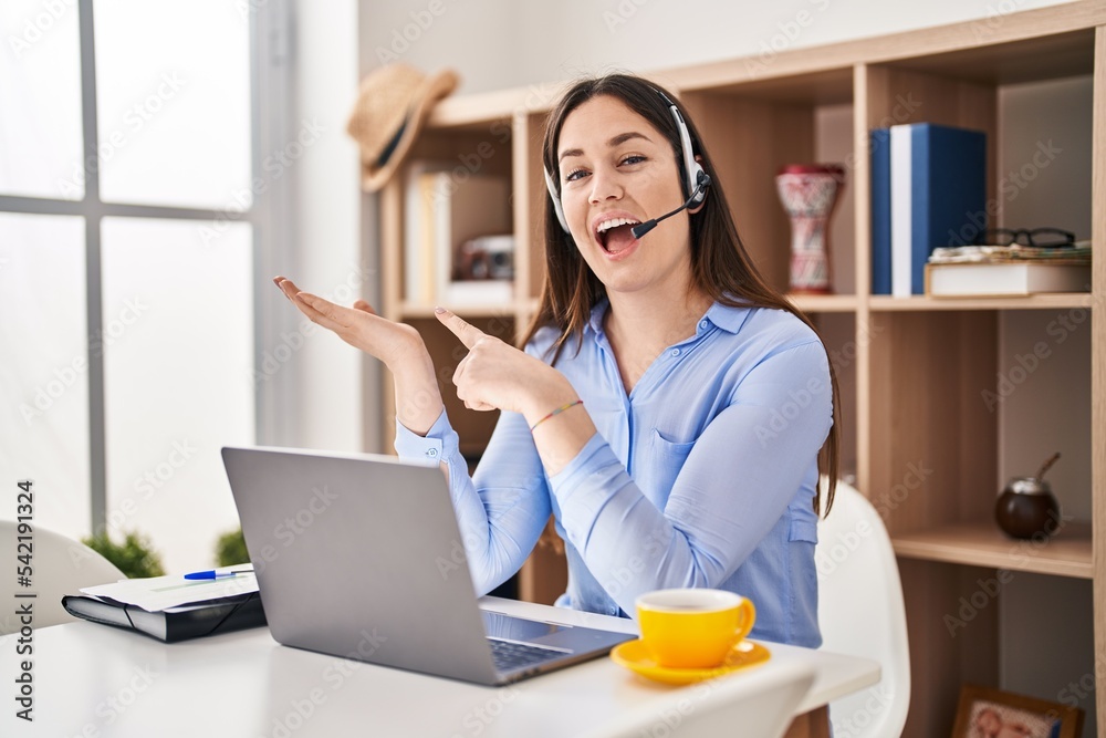Young brunette woman wearing call center agent headset amazed and smiling to the camera while presenting with hand and pointing with finger. Young brunette woman wearing call center agent headset amazed and smiling to the camera while presenting with hand and pointing with finger.