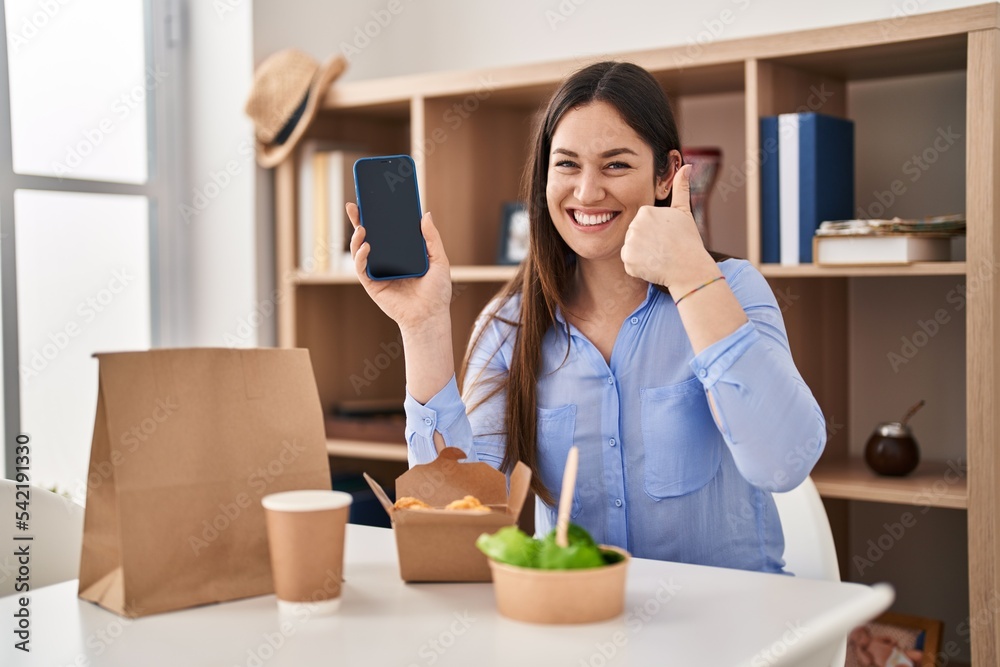Young brunette woman eating take away food at home showing smartphone screen smiling happy and positive, thumb up doing excellent and approval sign