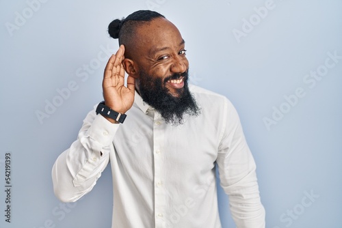 African american man standing over blue background smiling with hand over ear listening an hearing to rumor or gossip. deafness concept.