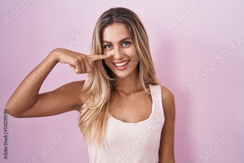 Young blonde woman standing over pink background pointing with hand finger to face and nose, smiling cheerful. beauty concept
