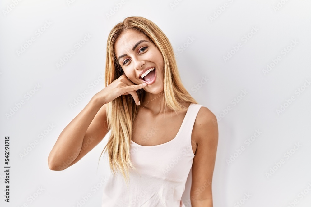 Blonde beautiful young woman standing over white isolated background smiling doing phone gesture with hand and fingers like talking on the telephone. communicating concepts. Blonde beautiful young woman standing over white isolated background smiling doing phone gesture with hand and fingers like talking on the telephone. communicating concepts.