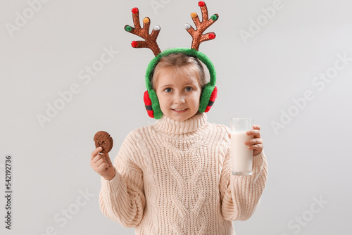 Happy little girl in reindeer horns with cookie and glass of milk on light background