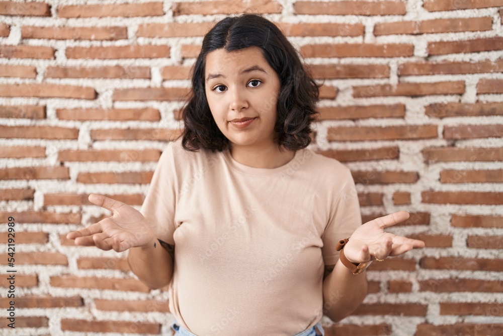 Young hispanic woman standing over bricks wall clueless and confused with open arms, no idea concept. Young hispanic woman standing over bricks wall clueless and confused with open arms, no idea concept.