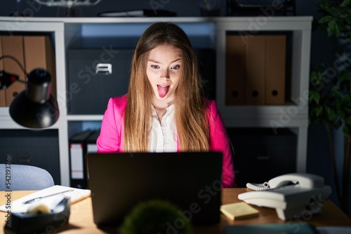 Young caucasian woman working at the office at night sticking tongue out happy with funny expression. emotion concept.