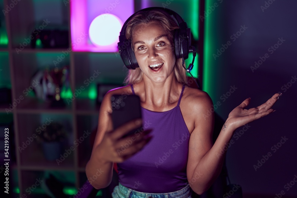 Young blonde woman playing video games with smartphone celebrating achievement with happy smile and winner expression with raised hand