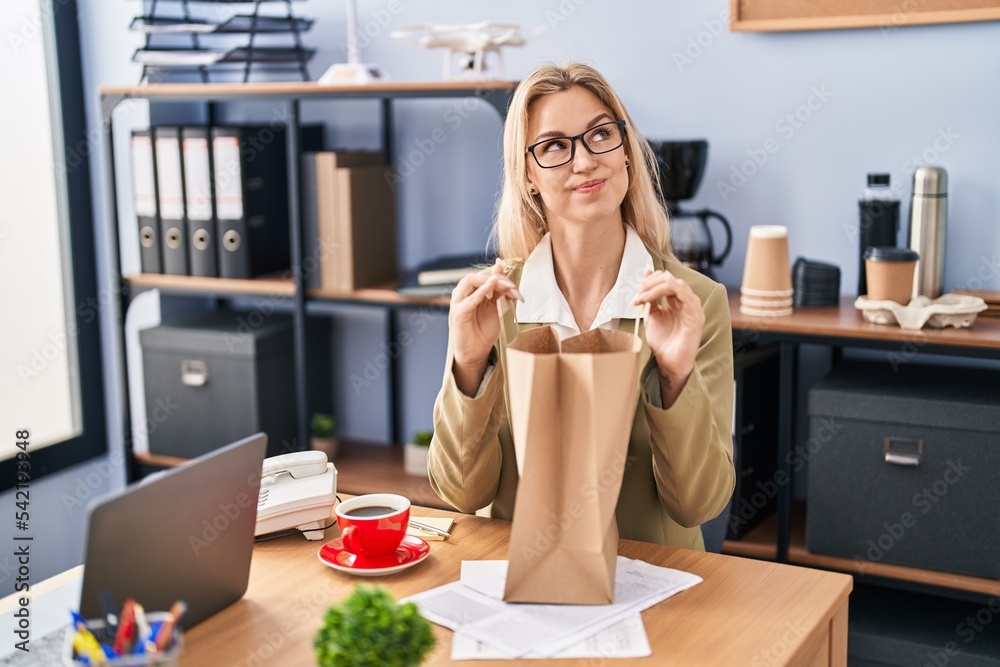 Young caucasian woman working at the office holding paper bag smiling looking to the side and staring away thinking.