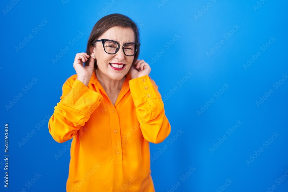 Middle age hispanic woman wearing glasses standing over blue background covering ears with fingers with annoyed expression for the noise of loud music. deaf concept. Middle age hispanic woman wearing glasses standing over blue background covering ears with fingers with annoyed expression for the noise of loud music. deaf concept.