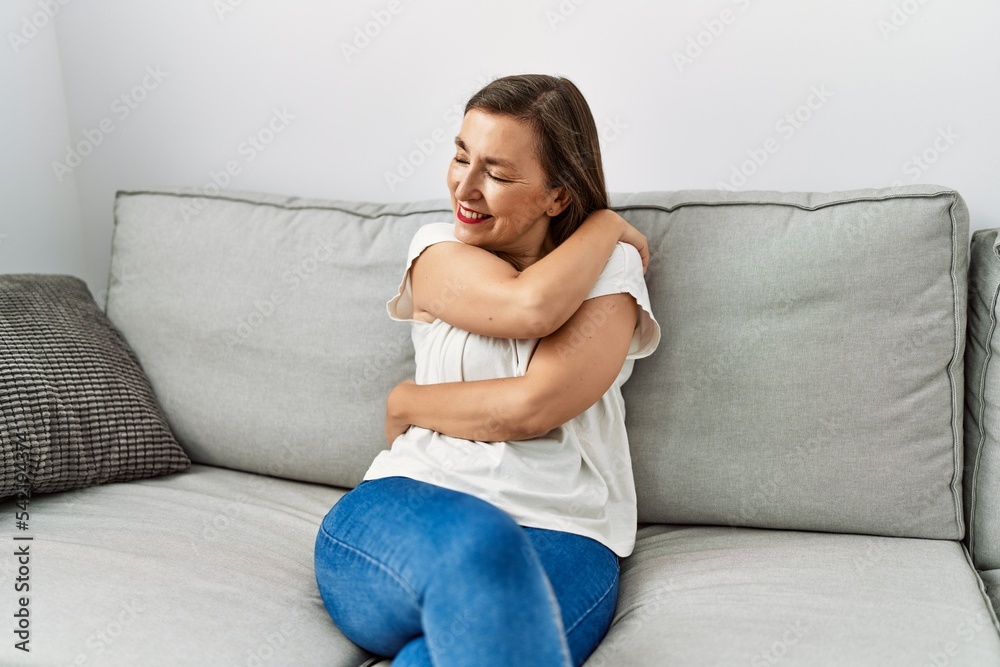 Middle age hispanic woman smiling sitting on the sofa hugging herself at home Middle age hispanic woman smiling sitting on the sofa hugging herself at home