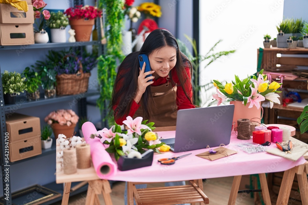 Young chinese woman florist talking on smartphone using laptop at flower shop