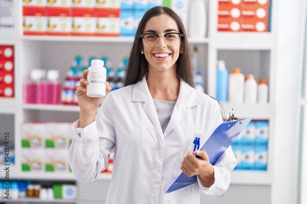Young brunette woman working at pharmacy drugstore holding pills smiling with a happy and cool smile on face. showing teeth.