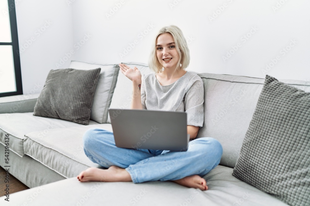 Young caucasian woman using laptop at home sitting on the sofa smiling cheerful presenting and pointing with palm of hand looking at the camera. Young caucasian woman using laptop at home sitting on the sofa smiling cheerful presenting and pointing with palm of hand looking at the camera.
