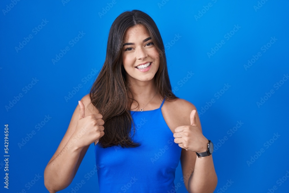 Hispanic woman standing over blue background success sign doing positive gesture with hand, thumbs up smiling and happy. cheerful expression and winner gesture. Hispanic woman standing over blue background success sign doing positive gesture with hand, thumbs up smiling and happy. cheerful expression and winner gesture.