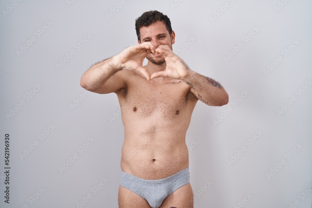 Young hispanic man standing shirtless wearing underware smiling in love doing heart symbol shape with hands. romantic concept.
