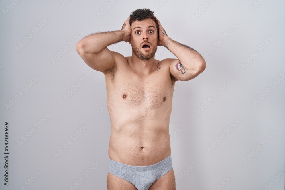 Young hispanic man standing shirtless wearing underware crazy and scared with hands on head, afraid and surprised of shock with open mouth