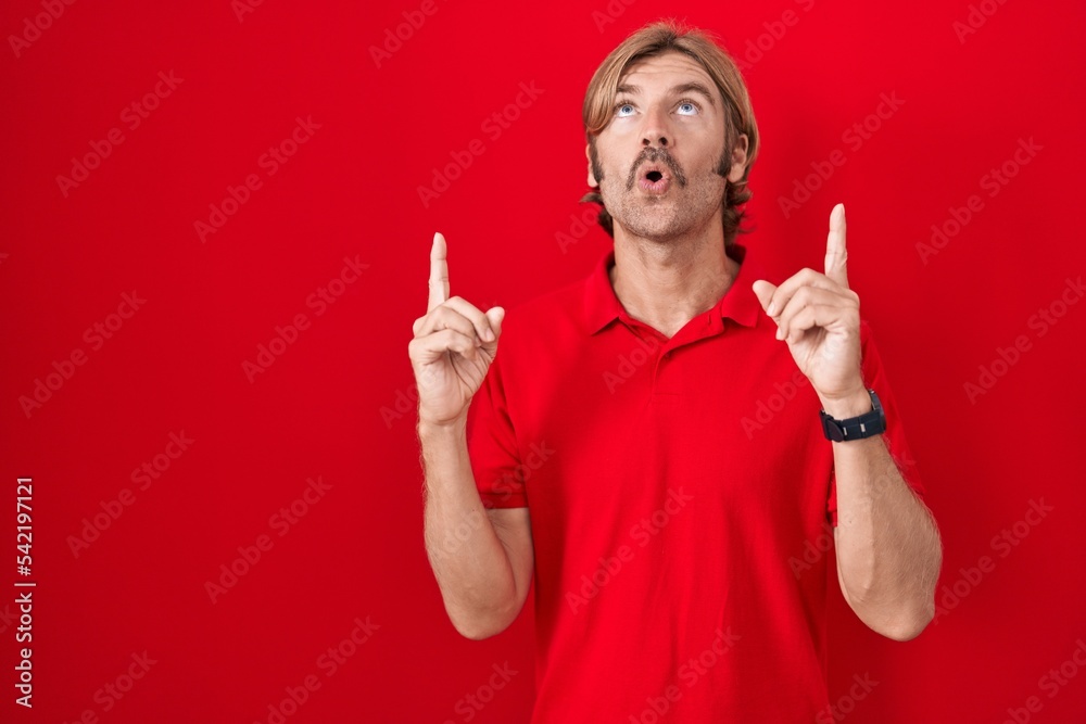 Caucasian man with mustache standing over red background amazed and surprised looking up and pointing with fingers and raised arms. Caucasian man with mustache standing over red background amazed and surprised looking up and pointing with fingers and raised arms.