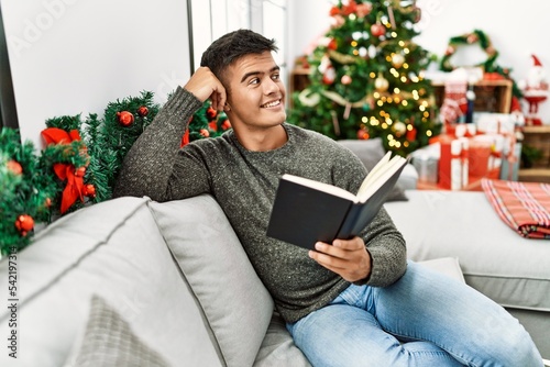 Young hispanic man reading book sitting on sofa by christmas tree at home © Krakenimages.com