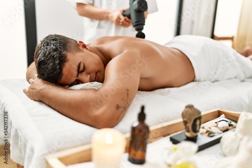 Young hispanic man relaxed having back massage using percussion pistol at beauty center