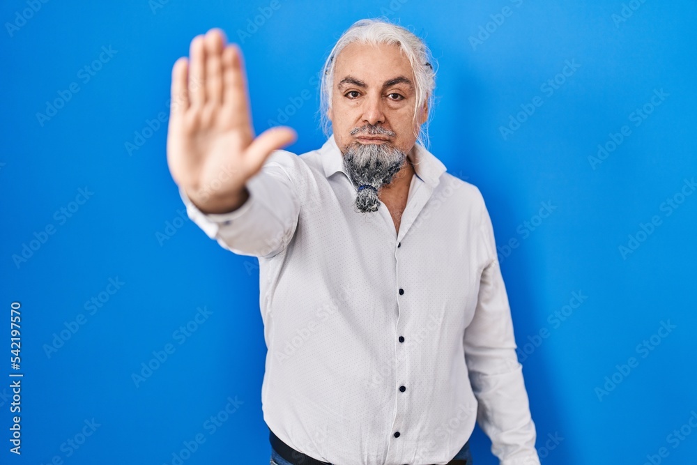 Middle age man with grey hair standing over blue background doing stop sing with palm of the hand. warning expression with negative and serious gesture on the face.