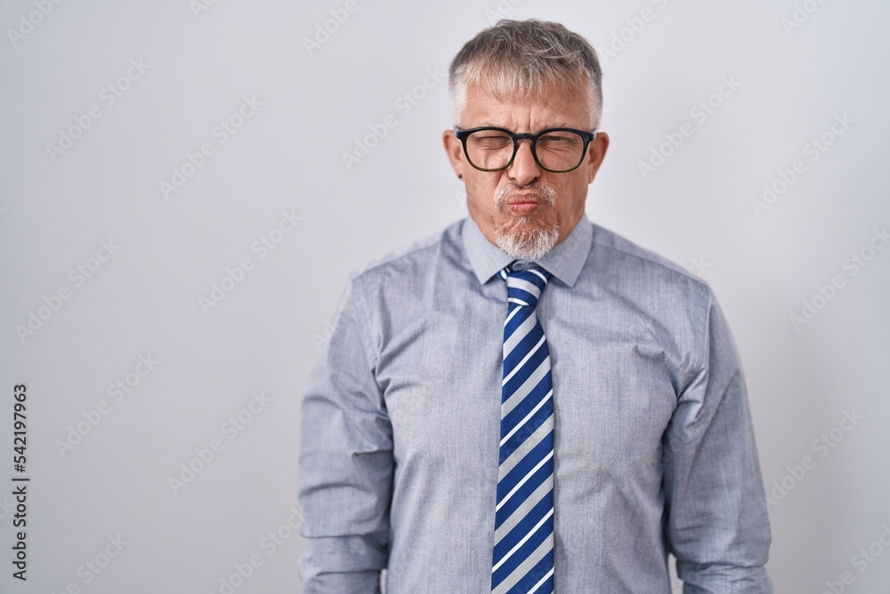 Hispanic business man with grey hair wearing glasses puffing cheeks with funny face. mouth inflated with air, crazy expression. Hispanic business man with grey hair wearing glasses puffing cheeks with funny face. mouth inflated with air, crazy expression.