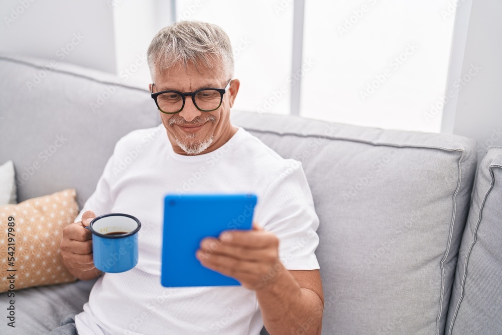 Middle age grey-haired man using touchpad drinking coffee at home Middle age grey-haired man using touchpad drinking coffee at home