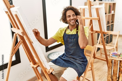 Young hispanic artist man smiling happy drawing and talking on the smartphone at art studio.