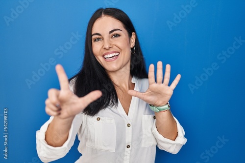 Young hispanic woman standing over blue background showing and pointing up with fingers number seven while smiling confident and happy.