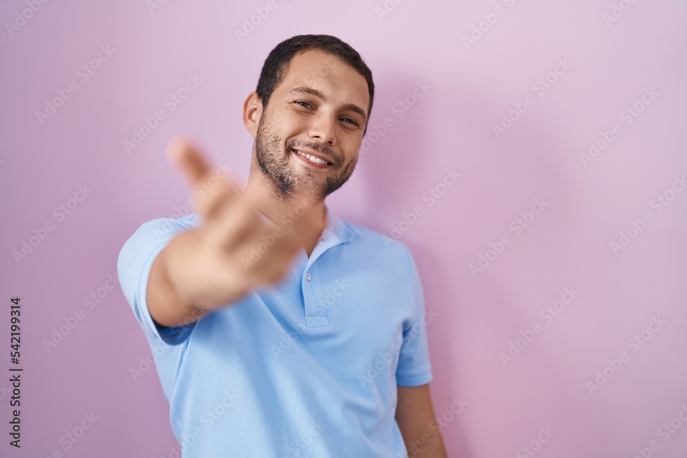 Hispanic man standing over pink background smiling cheerful offering palm hand giving assistance and acceptance. Hispanic man standing over pink background smiling cheerful offering palm hand giving assistance and acceptance.