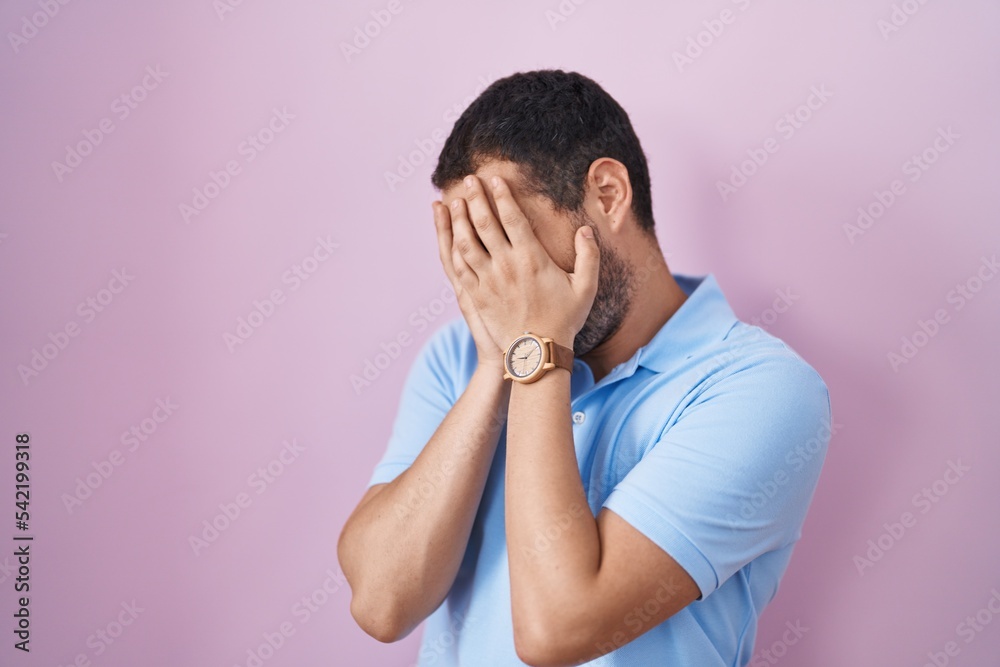 Hispanic man standing over pink background with sad expression covering face with hands while crying. depression concept. Hispanic man standing over pink background with sad expression covering face with hands while crying. depression concept.