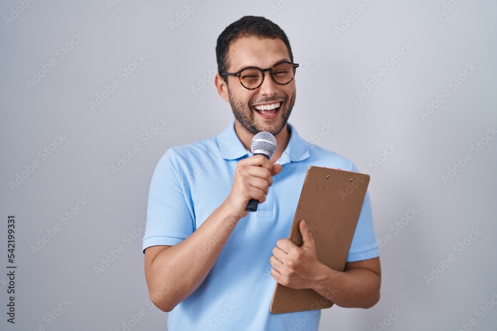 Hispanic man holding reporter microphone and clipboard winking looking at the camera with sexy expression, cheerful and happy face. Hispanic man holding reporter microphone and clipboard winking looking at the camera with sexy expression, cheerful and happy face.