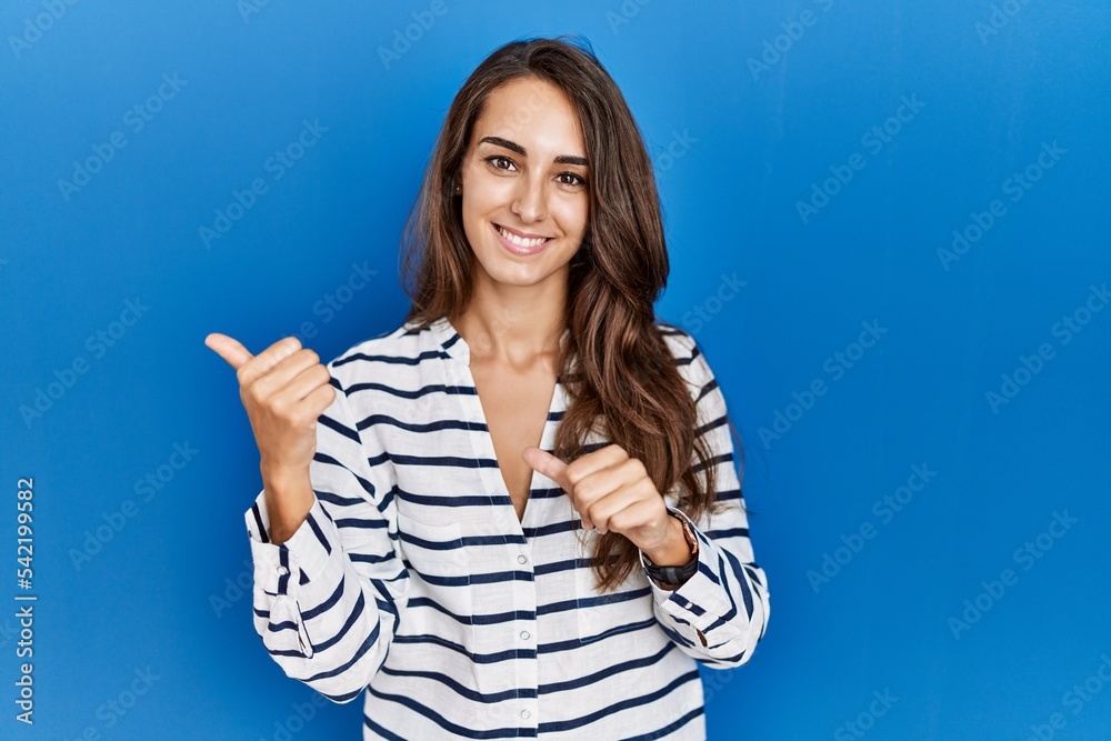 Young hispanic woman standing over blue isolated background pointing to the back behind with hand and thumbs up, smiling confident Young hispanic woman standing over blue isolated background pointing to the back behind with hand and thumbs up, smiling confident