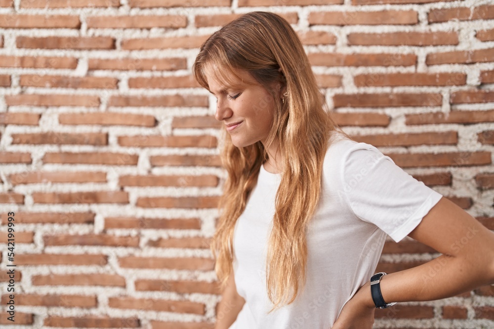 Young caucasian woman standing over bricks wall suffering of backache, touching back with hand, muscular pain