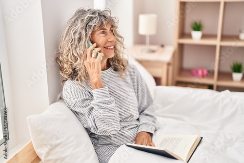 Middle age woman talking on the smartphone sitting on bed at bedroom