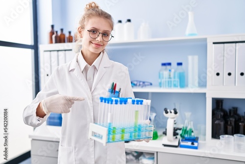 Young caucasian woman working at scientist laboratory holding samples smiling happy pointing with hand and finger