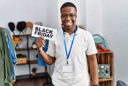 Young african man holding black friday banner at retail shop looking positive and happy standing and smiling with a confident smile showing teeth