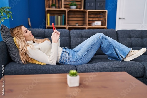 Young blonde woman using smartphone lying on sofa at home