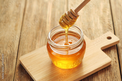 A transparent jar of honey stands on a wooden table. Honey flows into a jar on a honey stick