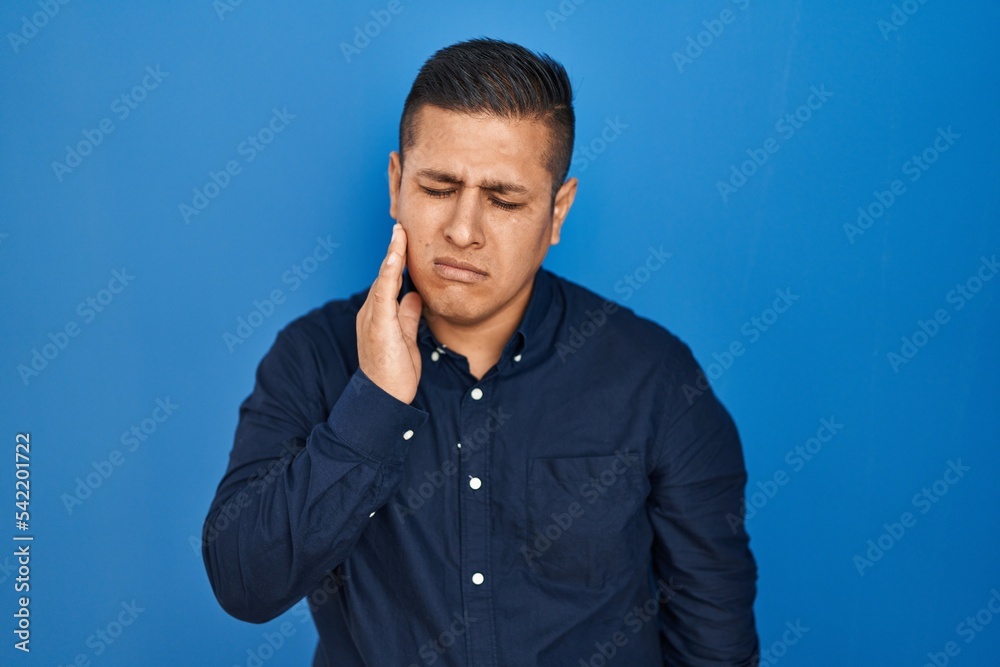 Hispanic young man standing over blue background touching mouth with hand with painful expression because of toothache or dental illness on teeth. dentist
