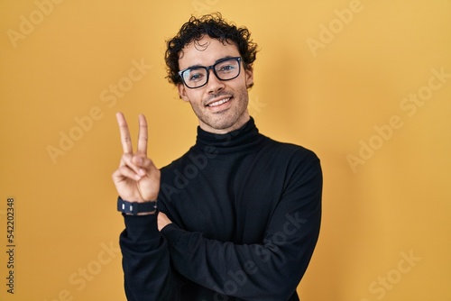 Hispanic man standing over yellow background smiling with happy face winking at the camera doing victory sign with fingers. number two.