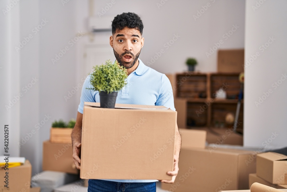 Hispanic man with beard moving to a new home holding box in shock face, looking skeptical and sarcastic, surprised with open mouth Hispanic man with beard moving to a new home holding box in shock face, looking skeptical and sarcastic, surprised with open mouth
