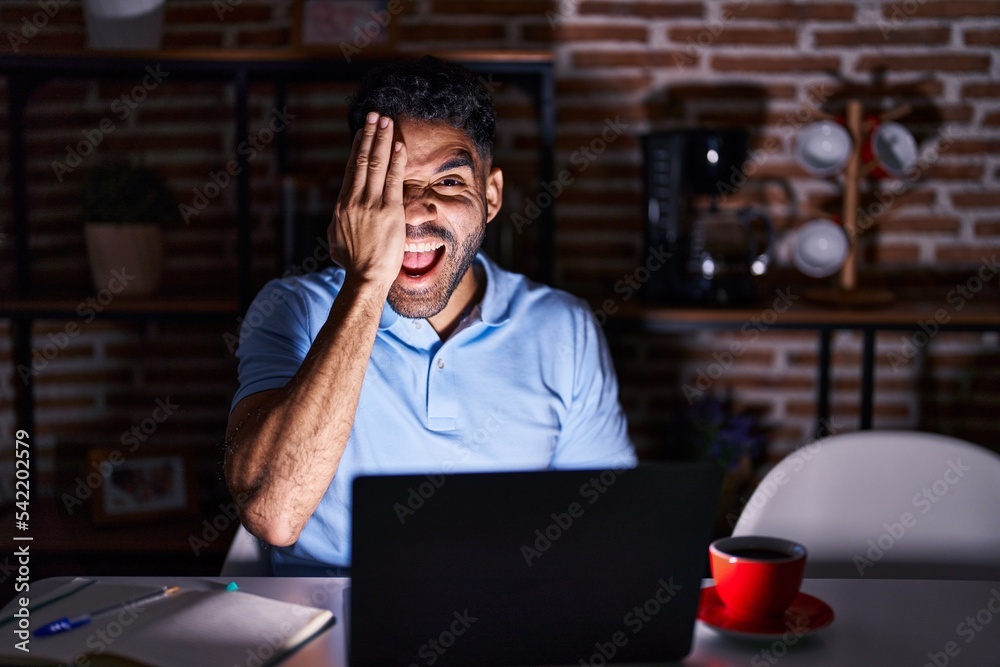 Hispanic man with beard using laptop at night covering one eye with hand, confident smile on face and surprise emotion. Hispanic man with beard using laptop at night covering one eye with hand, confident smile on face and surprise emotion.