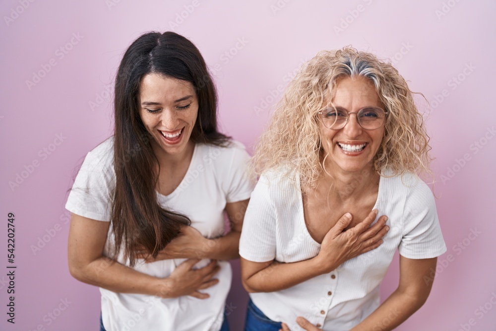 Mother and daughter standing together over pink background smiling and laughing hard out loud because funny crazy joke with hands on body. Mother and daughter standing together over pink background smiling and laughing hard out loud because funny crazy joke with hands on body.