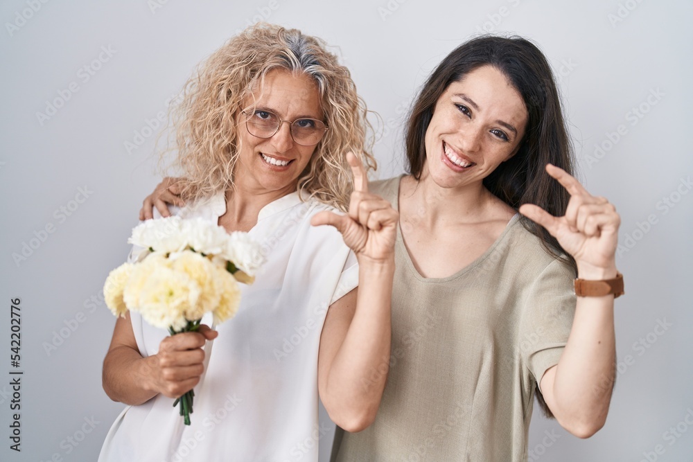 Mother and daughter holding bouquet of white flowers smiling and confident gesturing with hand doing small size sign with fingers looking and the camera. measure concept. Mother and daughter holding bouquet of white flowers smiling and confident gesturing with hand doing small size sign with fingers looking and the camera. measure concept.