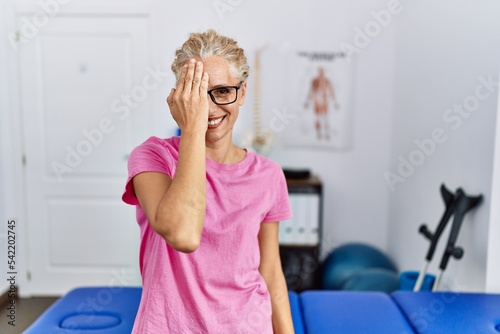 Middle age blonde woman at pain recovery clinic covering one eye with hand  confident smile on face and surprise emotion.