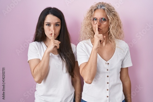 Mother and daughter standing together over pink background asking to be quiet with finger on lips. silence and secret concept.