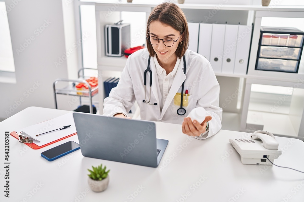 Young caucasian woman doctor using laptop holding pills at clinic Young caucasian woman doctor using laptop holding pills at clinic