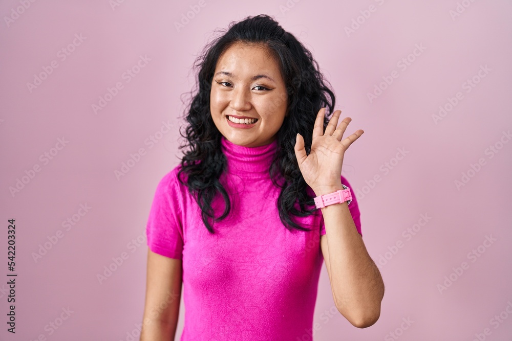 Young asian woman standing over pink background waiving saying hello happy and smiling, friendly welcome gesture Young asian woman standing over pink background waiving saying hello happy and smiling, friendly welcome gesture