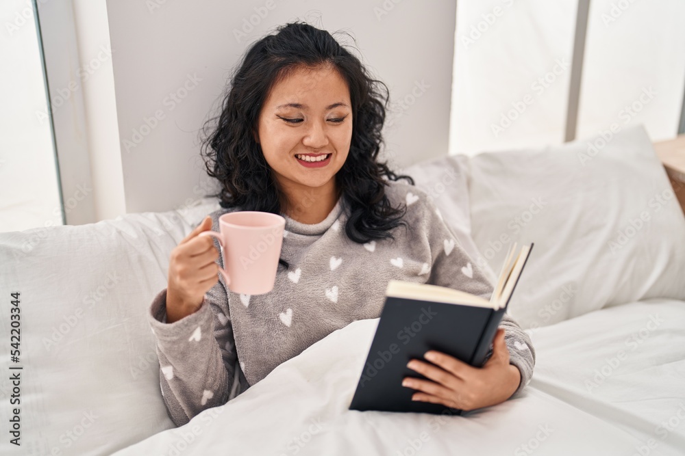 Young chinese woman dinking coffee and reading book sitting on bed at bedroom Young chinese woman dinking coffee and reading book sitting on bed at bedroom