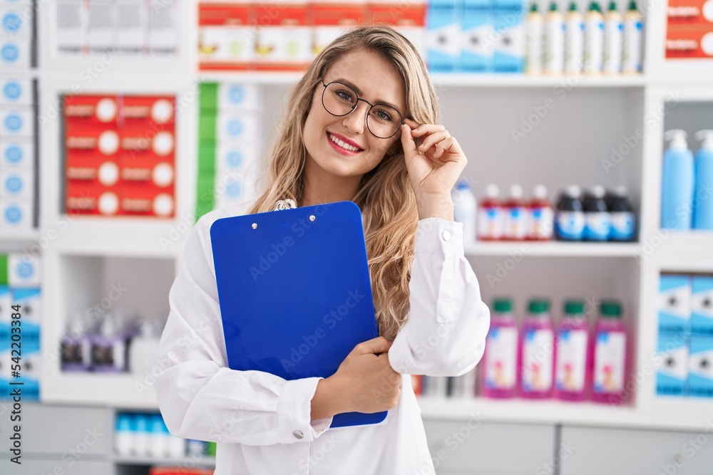 Young woman pharmacist smiling confident holding clipboard at pharmacy